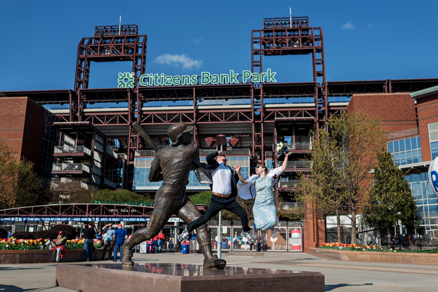 A bride and groom jump while holding hands in front of Citizens Bank Park in Philadelphia.